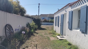 Un camí estret que condueix a un edifici amb persianes blaves, envoltat per una paret blanca i diferents plantes.