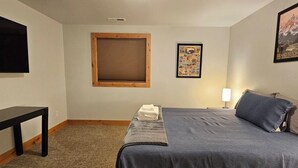 Ein Schlafzimmer mit einem Bett, einem Nachttisch, einem Fenster mit Jalousien, einem an der Wand montierten Fernseher und einem gerahmten Poster.