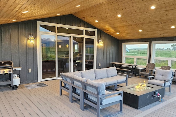 A covered outdoor area with a wooden deck, a sofa, chairs, a barbecue grill, and a fire pit. - Alpenglo Farm with Event Space and Parking (Driggs)