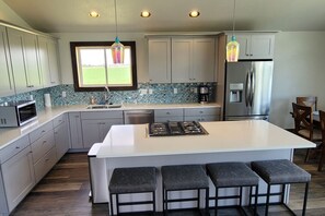 A modern kitchen with a central island, bar stools, a stainless steel refrigerator, and a window with a view of a green field. - Alpenglo Farm with Event Space and Parking (Driggs)