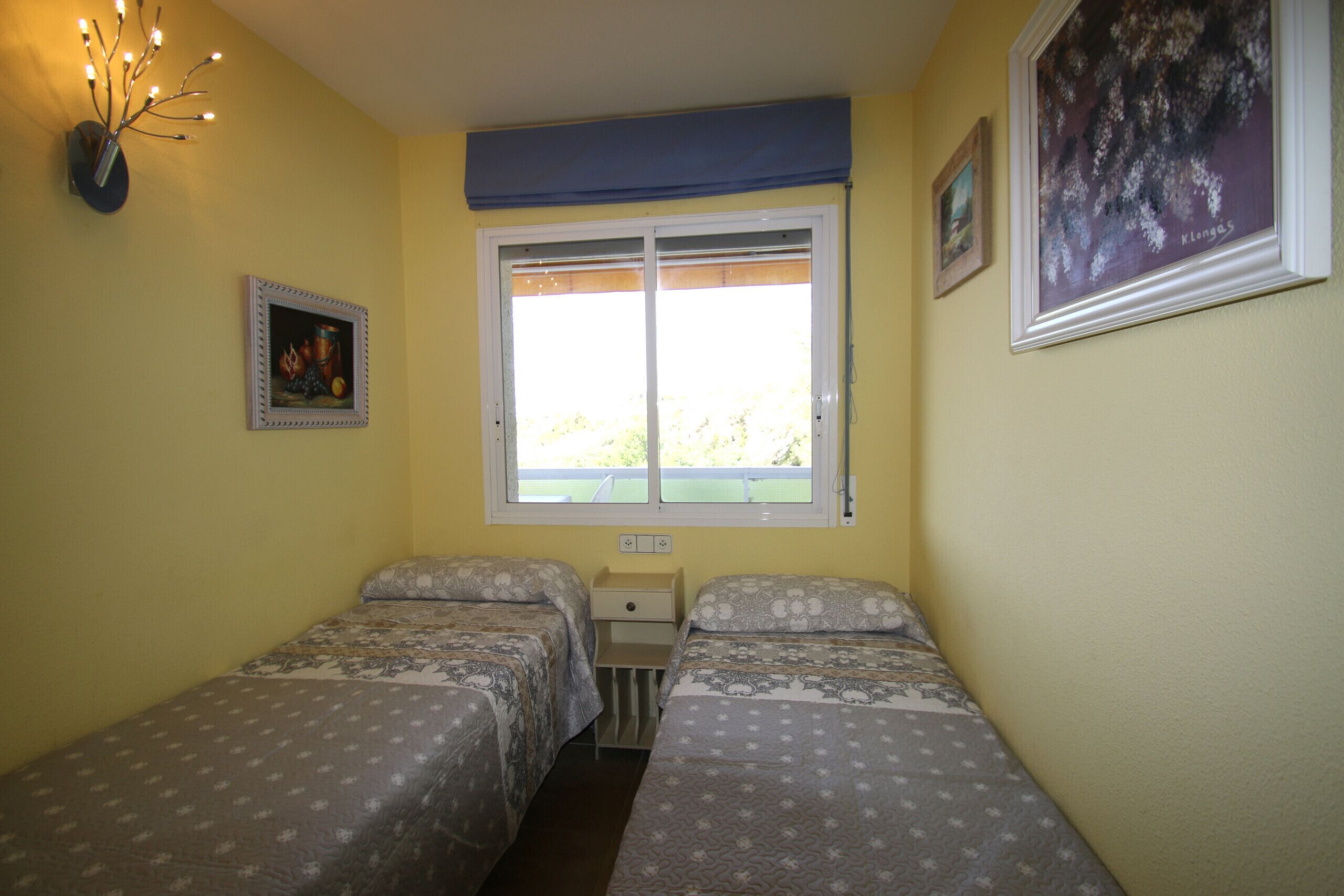 A bedroom with two beds, a window with a view, and a wall-mounted light fixture.