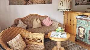 Una sala de estar acogedora con un sofá a rayas, un sillón de mimbre, una pequeña mesa redonda con mantel floral y un aparador con paneles decorativos.