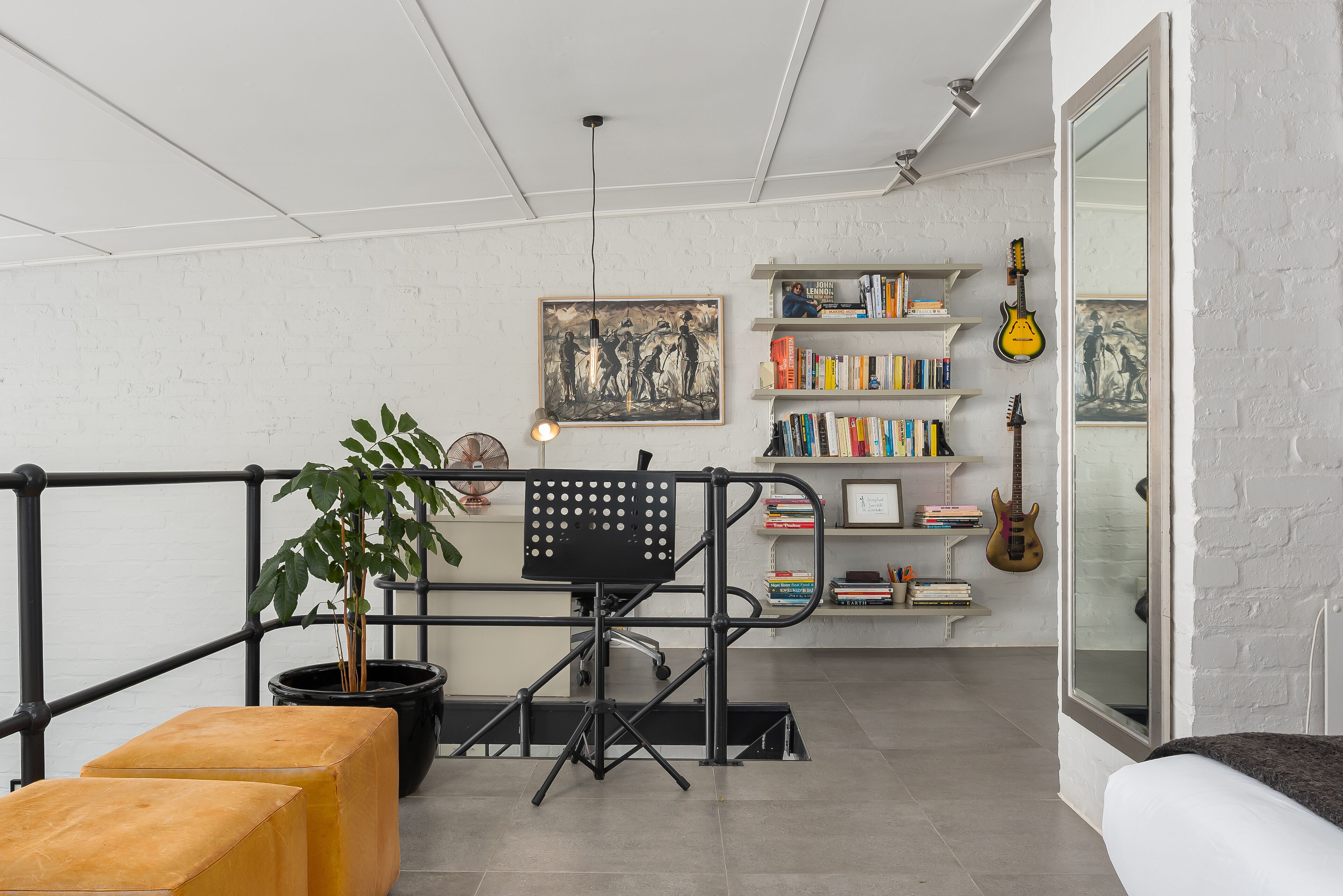 Ein Zimmer mit weißer Ziegelwand, einem schwarzen Metaltschreibtisch, einem Topfpflanzen und einem Bücherregal mit Büchern und Gitarren.