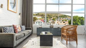 Ein modernes Wohnzimmer mit einem grauen Sofa, einem schwarzen Couchtisch und einem Korbstuhl. Ein großes Fenster bietet Blick auf eine Hügelstadt.