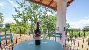 Un tavolo rotondo con una bottiglia di vino e due bicchieri su un balcone, vista su alberi e cielo sereno.