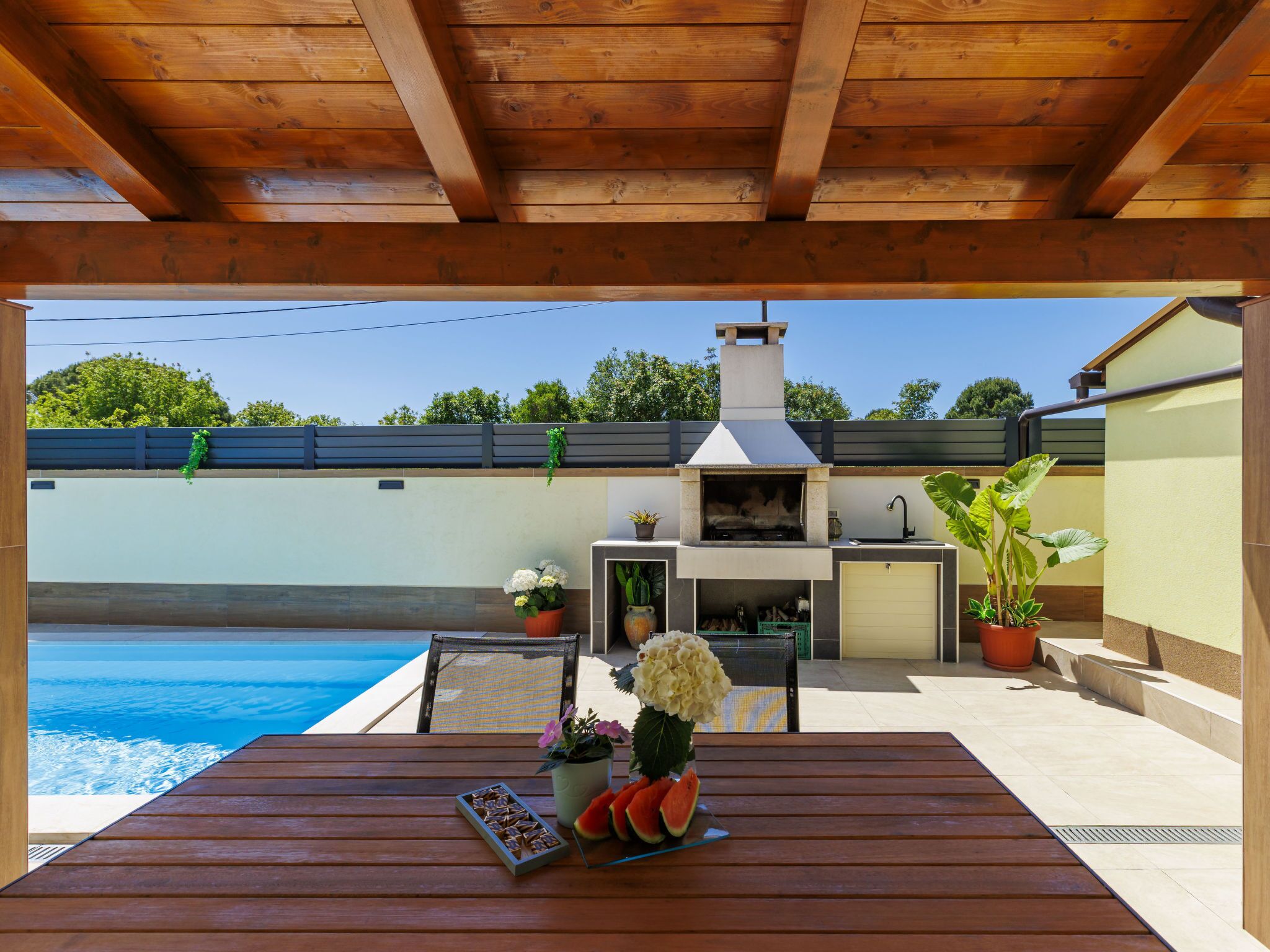 An outdoor patio with a wooden table, a barbecue grill, and a pool.