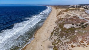 An aerial view of a coastal area with a sandy beach, a road curving along the coast, and sparse vegetation.