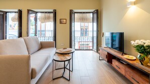 A modern living room with a sofa, a small round table, a TV on a wooden stand, and a vase with flowers.
