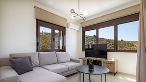 A modern living room with a sofa, a TV, a small table, and large windows offering a view of a hilly landscape. - Villa Lia with private ecologic pool and amazing view! (Kontopoula)