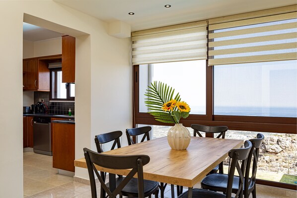 A dining area with a wooden table and chairs, a vase with flowers, and a view of the sea through large windows.