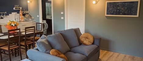 A modern living room with a grey sofa, a dining table with chairs, a coffee table with a remote control, and a painting on the wall.