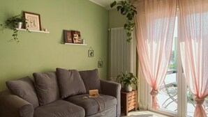 A living room with a sofa, a hanging plant, a radiator, and a window with sheer curtains. - WellHome   "Ruchè" Apartment (ASTI)