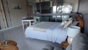 A modern bedroom with a bed, a sofa, a dining table, and a kitchen area visible through a glass door.