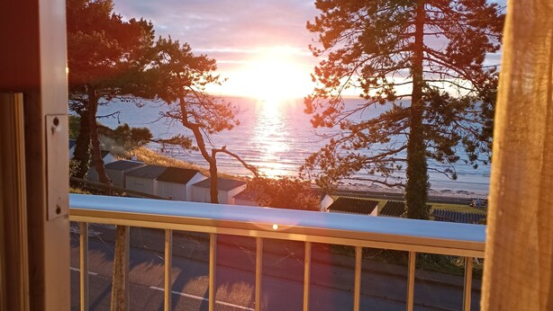A view from a balcony overlooking a sunset over the ocean, with trees and a building in the background.