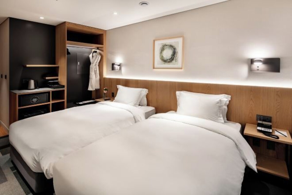 A hotel room with two beds, a wardrobe, a small desk, and a wall-mounted light fixture.