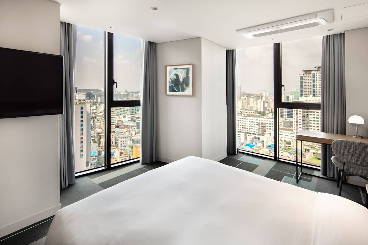 Habitación de hotel con una cama grande, un escritorio y una silla. Ofrece vistas a la ciudad a través de amplias ventanas.