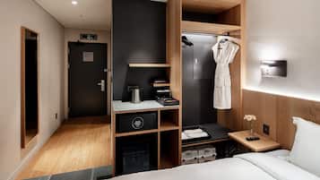 A modern hotel room with a bed, a wooden wardrobe, a small desk with a coffee maker, a robe hanging, and a nightstand with a vase of flowers.