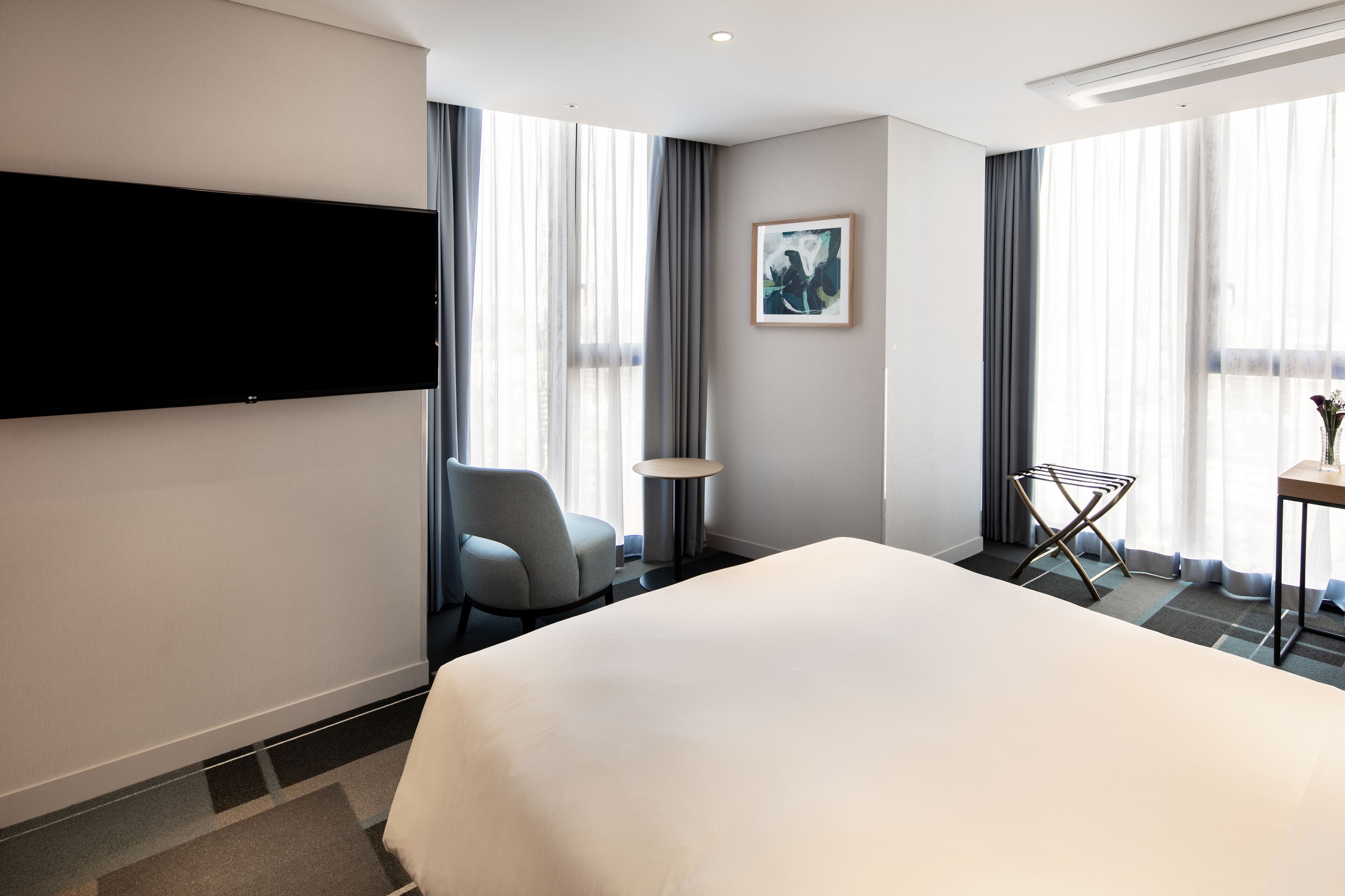 A modern hotel room with a large bed, a flat-screen TV mounted on the wall, a chair, a small round table, and a window with sheer curtains.