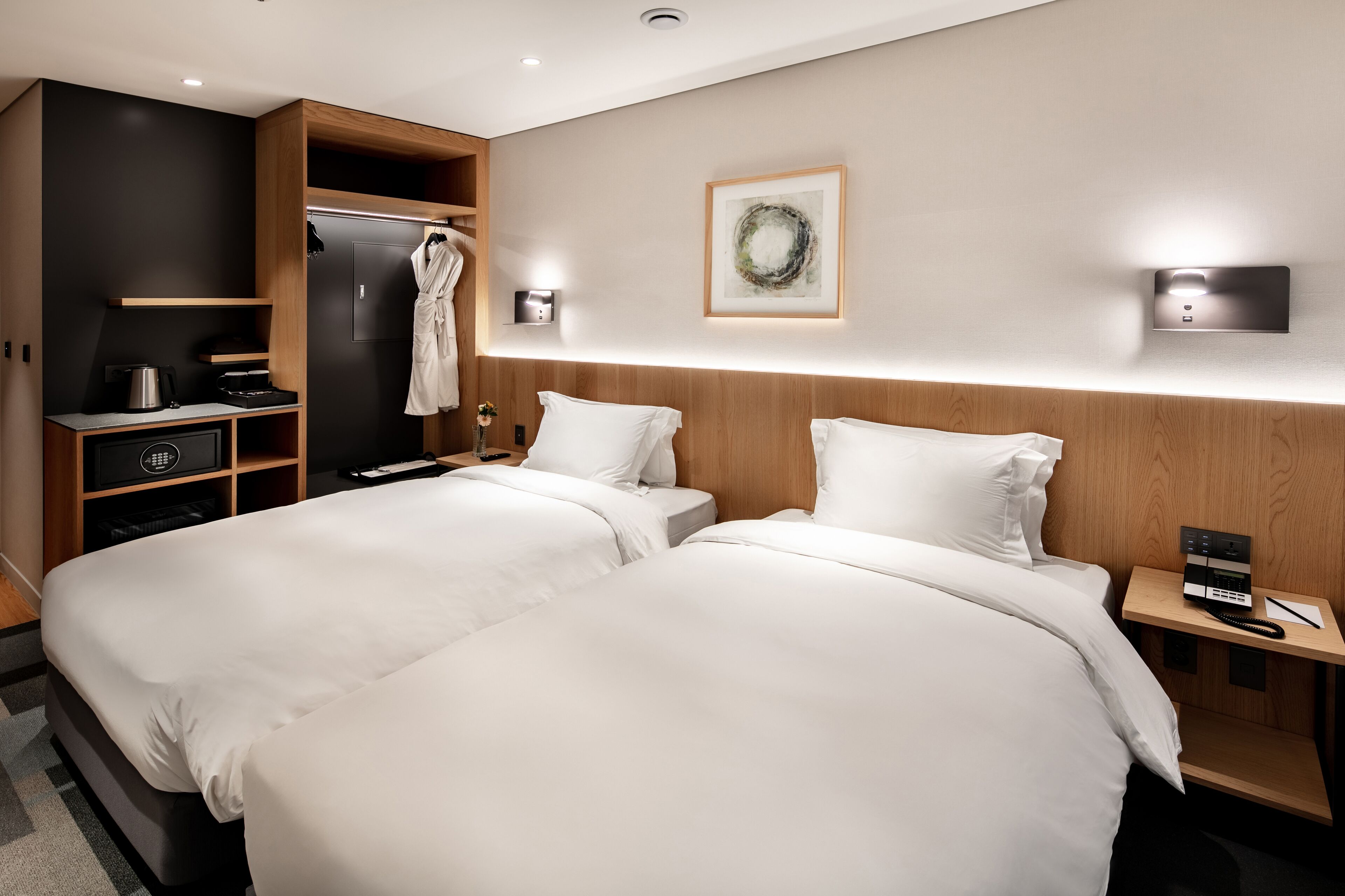 A hotel room with two beds, a robe closet, a coffee machine, and a wall-mounted light fixture.
