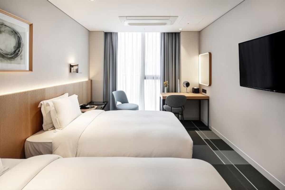 A hotel room with two beds, a desk, a chair, a TV, and a large window with curtains.