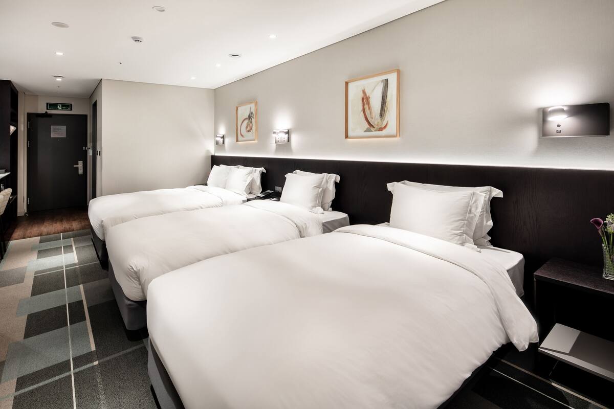 Una habitación de hotel con tres camas individuales, un cabecero oscuro y una mesita de noche pequeña con un florero.