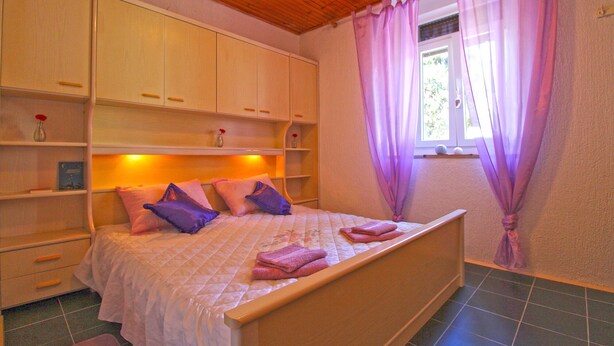 A bedroom with a bed, wooden headboard, built-in shelves, a window with purple curtains, and a tiled floor. - Apartment 2192/56814 (Istria - Pula), 3000m from the beach (Pula)