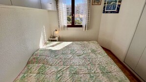 A small bedroom with a bed, a nightstand, a window with curtains, and a wall-mounted shelf.
