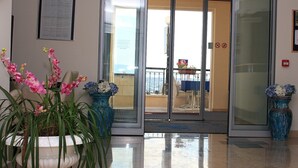 Et hotel lobby med en glasdør, der fører ud til en balkon med udendørs siddepladser. Der står plantekasser med blomster på gulvet.