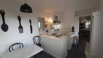 Une cuisine compacte avec un plan de travail blanc, une table à manger et des chaises. On y trouve des ustensiles de cuisine accrochés au mur, un four à micro-ondes et une machine à café sur le plan de travail.