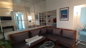 Un salon d’angle avec un canapé marron, un plaid blanc, une table basse en verre et un ventilateur blanc. On y trouve également une étagère blanche ornée d’objets décoratifs et un tableau encadré accroché au mur.