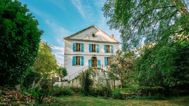 A two-story white building with blue shutters, surrounded by lush greenery and trees. - Le Moulin du Pont (Mezin)