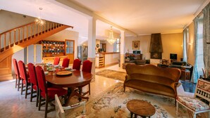 A spacious living area with a wooden staircase, a dining table with red chairs, a sofa, and a fireplace. - Le Moulin du Pont (Mezin)