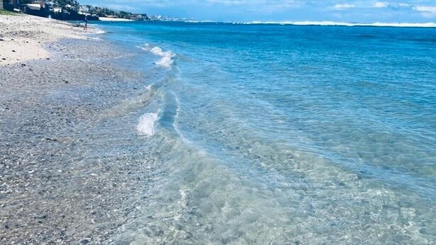 Una playa con aguas cristalinas y poco profundas, una orilla de arena y edificios al fondo.