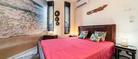 A bedroom with a large bed, a wooden headboard, a nightstand, a wall-mounted air conditioner, and a beach mural.