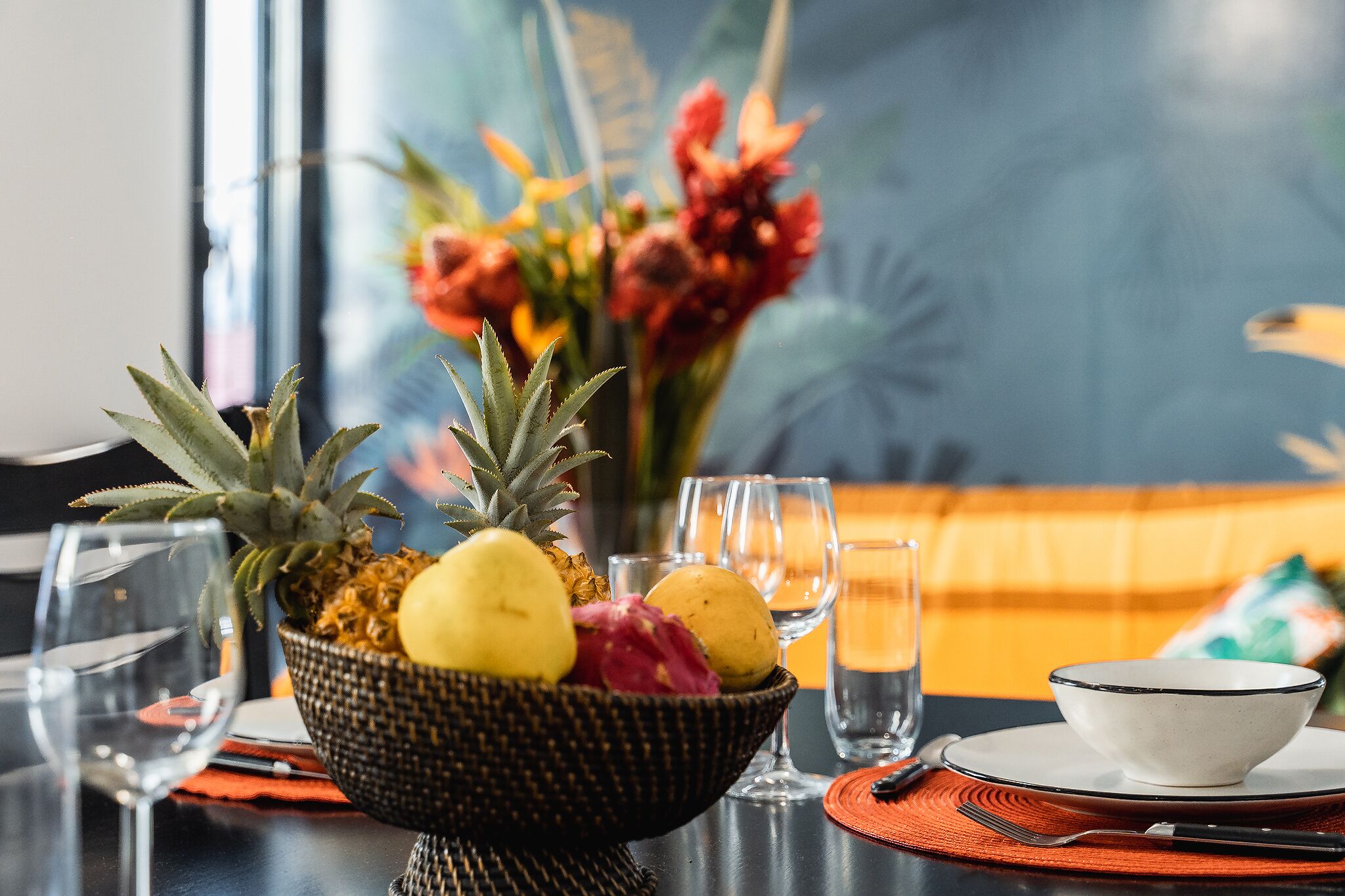 Une table à manger avec un panier de fruits, des verres et un bol blanc posé sur un set de table.