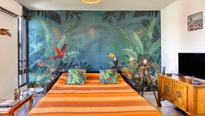 Un dormitorio con cama, papel tapiz con motivos tropicales, televisor y un mueble de madera.