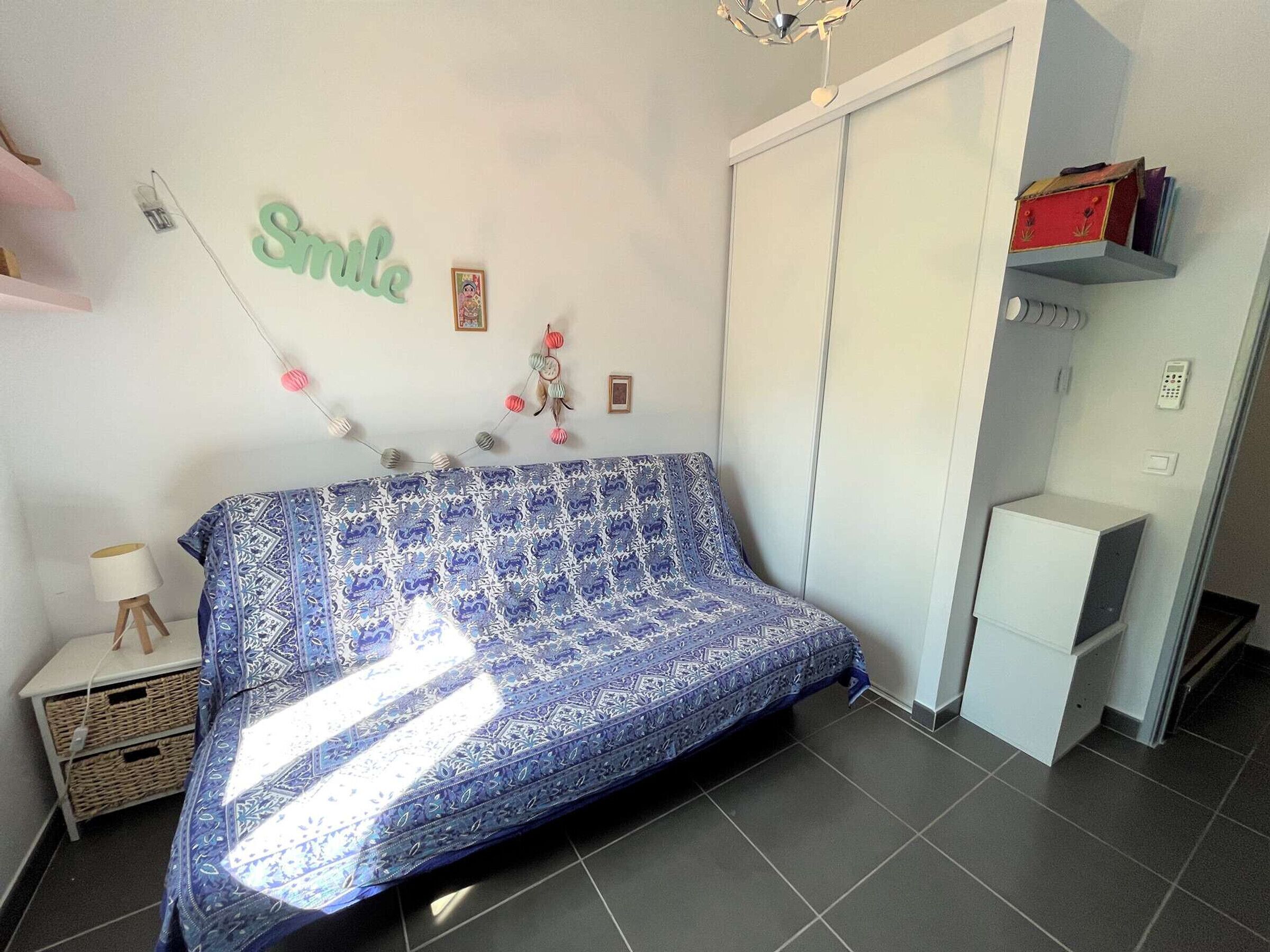 Un dormitorio con una colcha estampada, un armario, una mesita de noche con lámpara y un estante en la pared con libros.