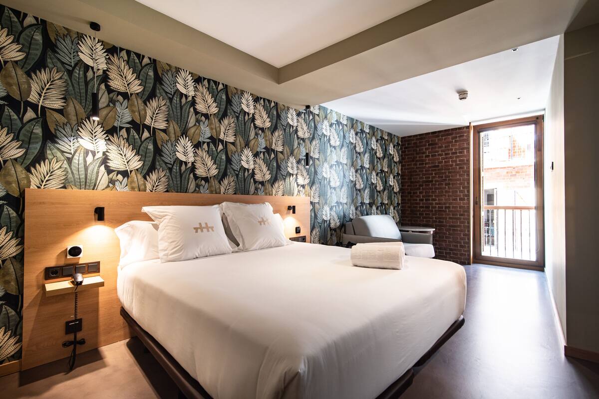 A hotel room with a large bed, a wooden headboard, a wall with a leafy pattern, a brick wall, and a sliding glass door to the outside.