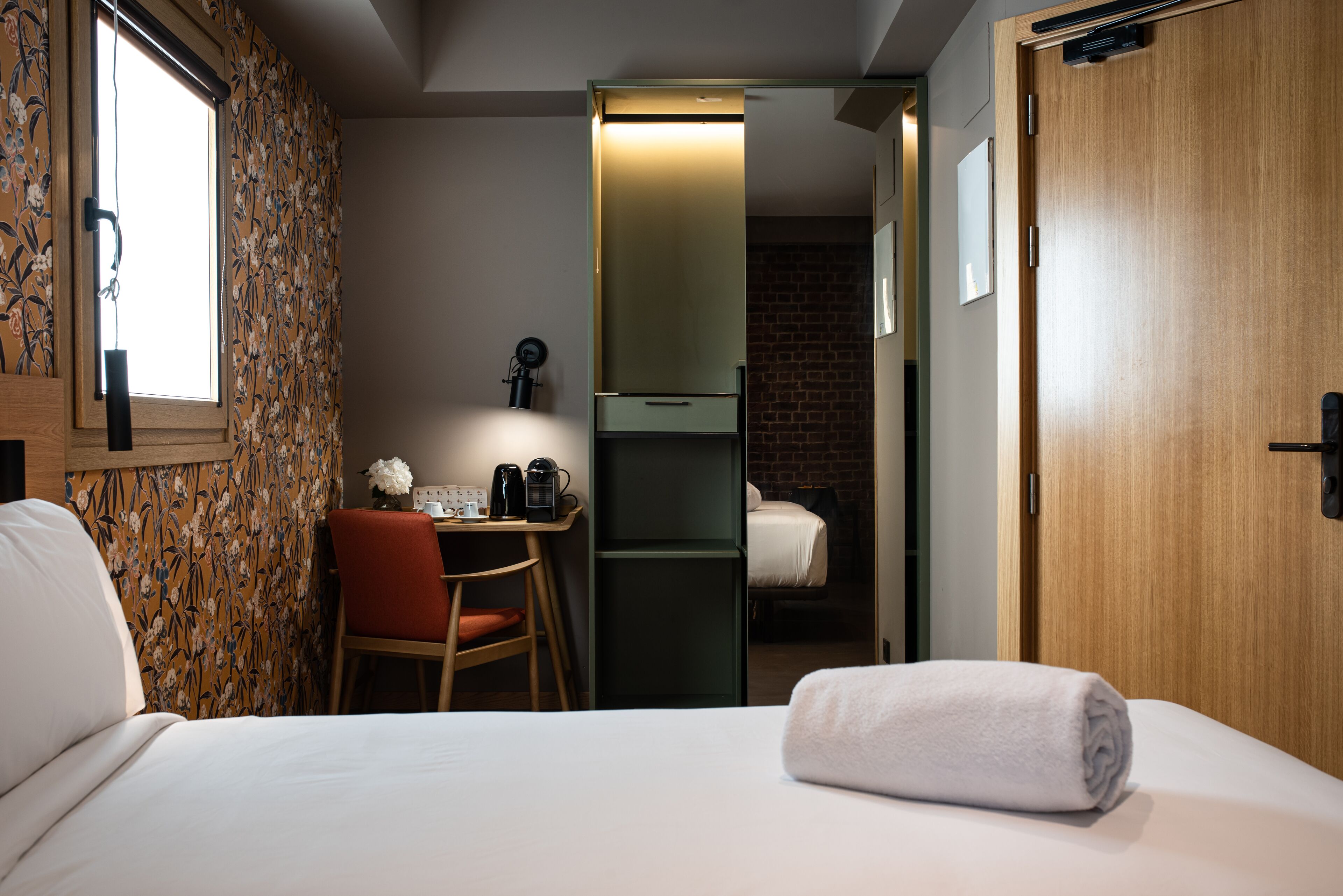 A compact hotel room with a bed, a desk with a chair, a small table with a vase, and a visible door leading to another room.