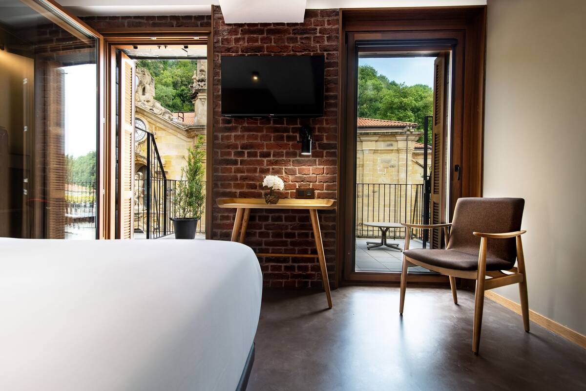 A hotel room with a bed, a TV mounted on a brick wall, a wooden chair, a small table with a vase, and a balcony with a view of greenery and buildings.