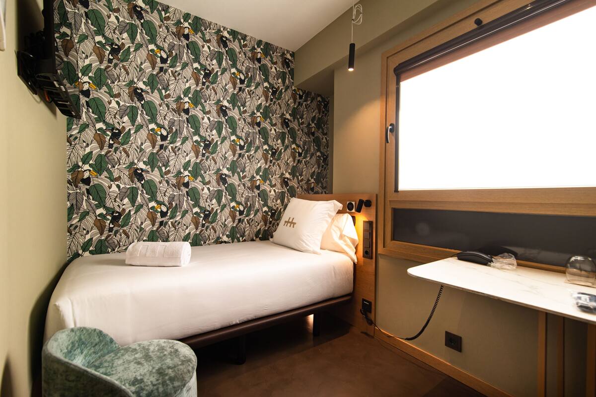 A hotel room with a bed, a bedside table, a chair, and a large window.