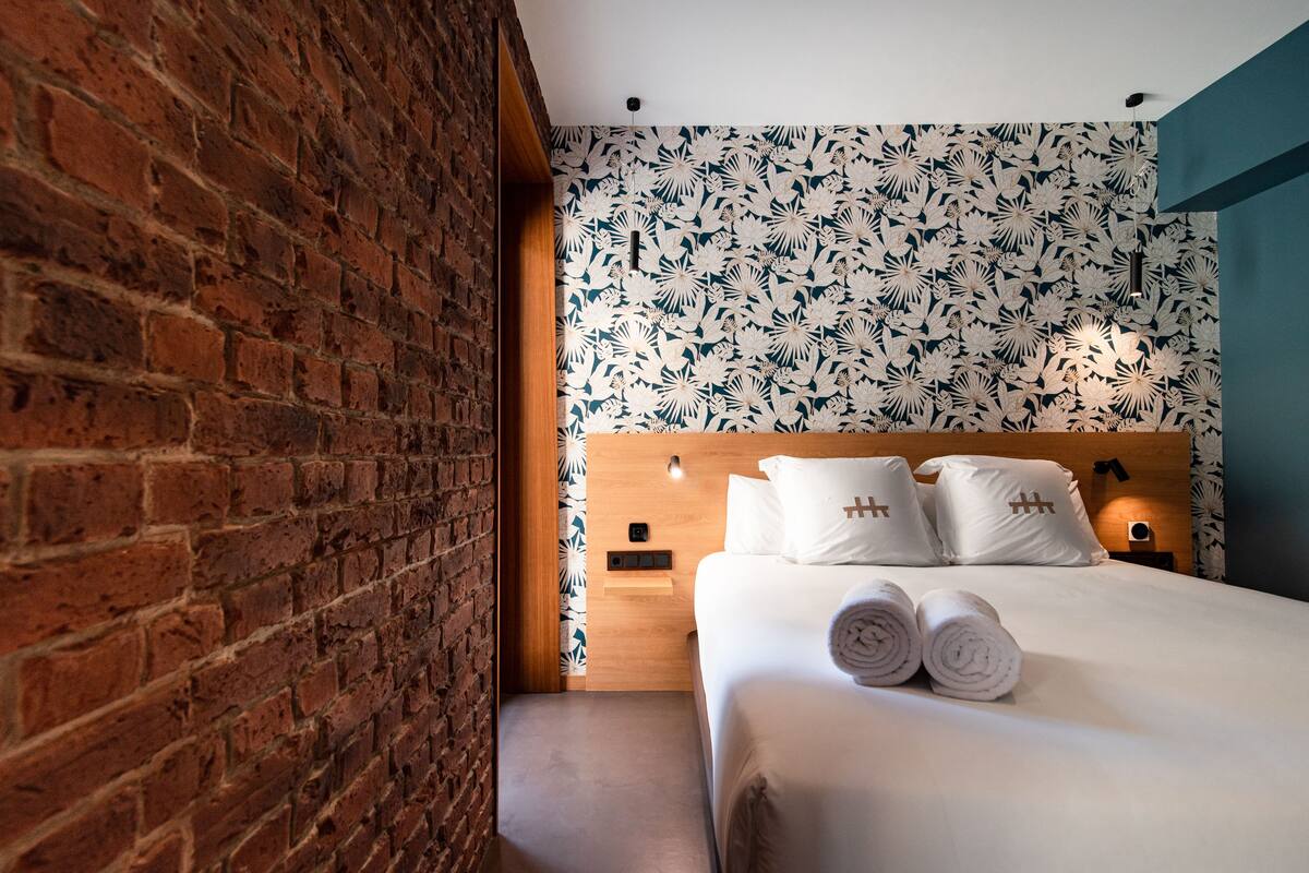 A hotel room with a brick wall, a wooden headboard, a floral wallpaper, and a neatly made bed with towels.