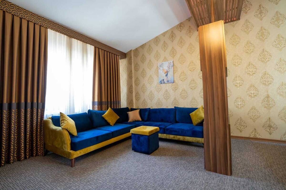 A room with a blue sofa, yellow cushions, a wooden coffee table, patterned wallpaper, and curtains.