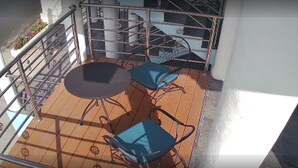 Un balcon avec une table et deux chaises, une rambarde en métal et un escalier en arrière-plan.