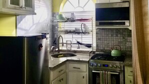 Une cuisine compacte avec des meubles blancs, un réfrigérateur en acier inoxydable, un évier avec un égouttoir et une cuisinière avec une bouilloire.