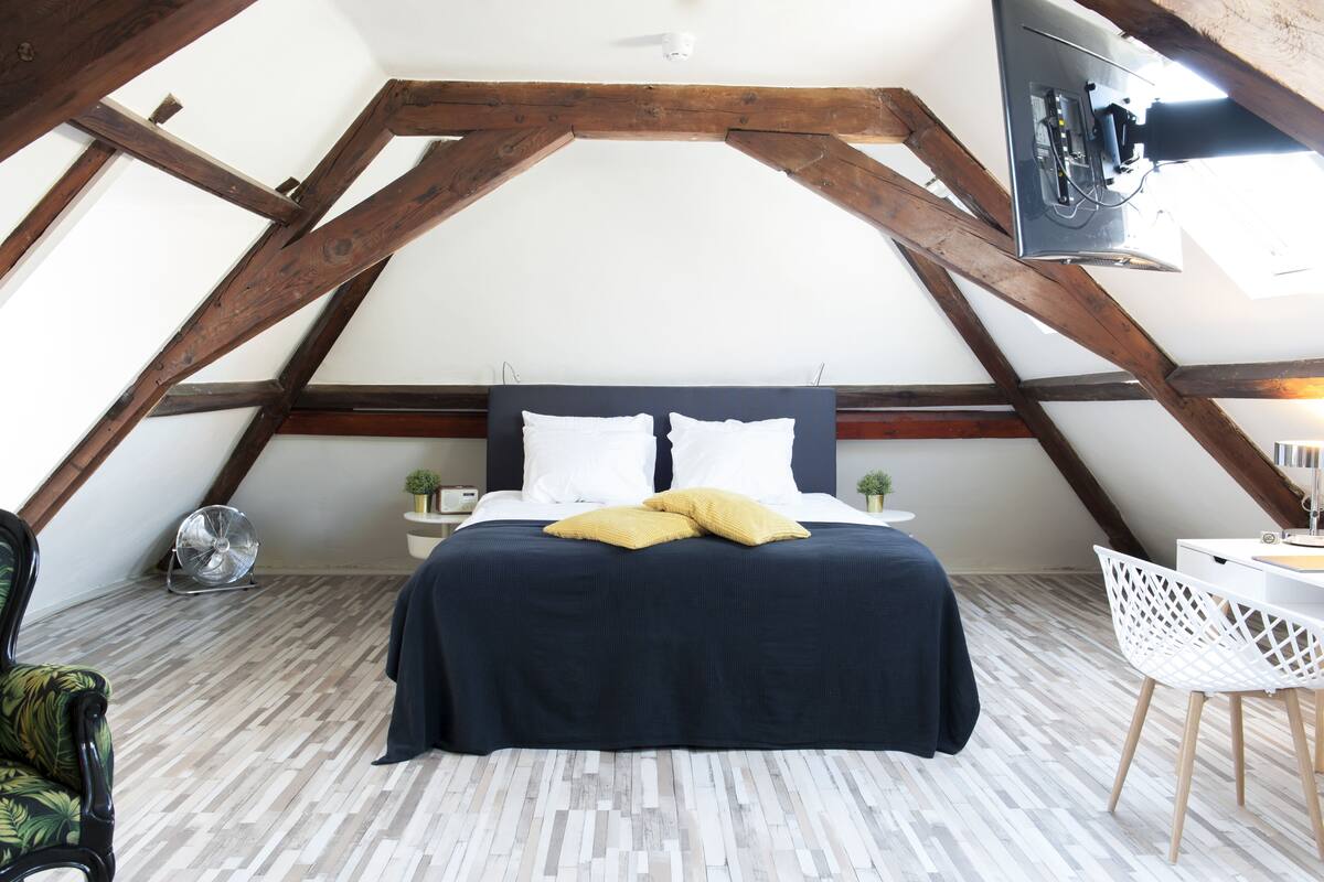 Ein Schlafzimmer mit schrägen Wänden, einem Bett mit dunkelblauer Bettdecke und weißen Kissen, einem kleinen Nachttisch mit Pflanze und einem weißen Stuhl.