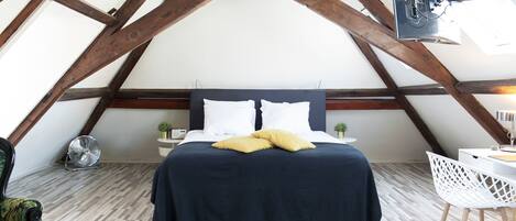 A bedroom with a sloped ceiling, a bed with a dark blue bedspread and white pillows, a small bedside table with a plant, and a white chair.
