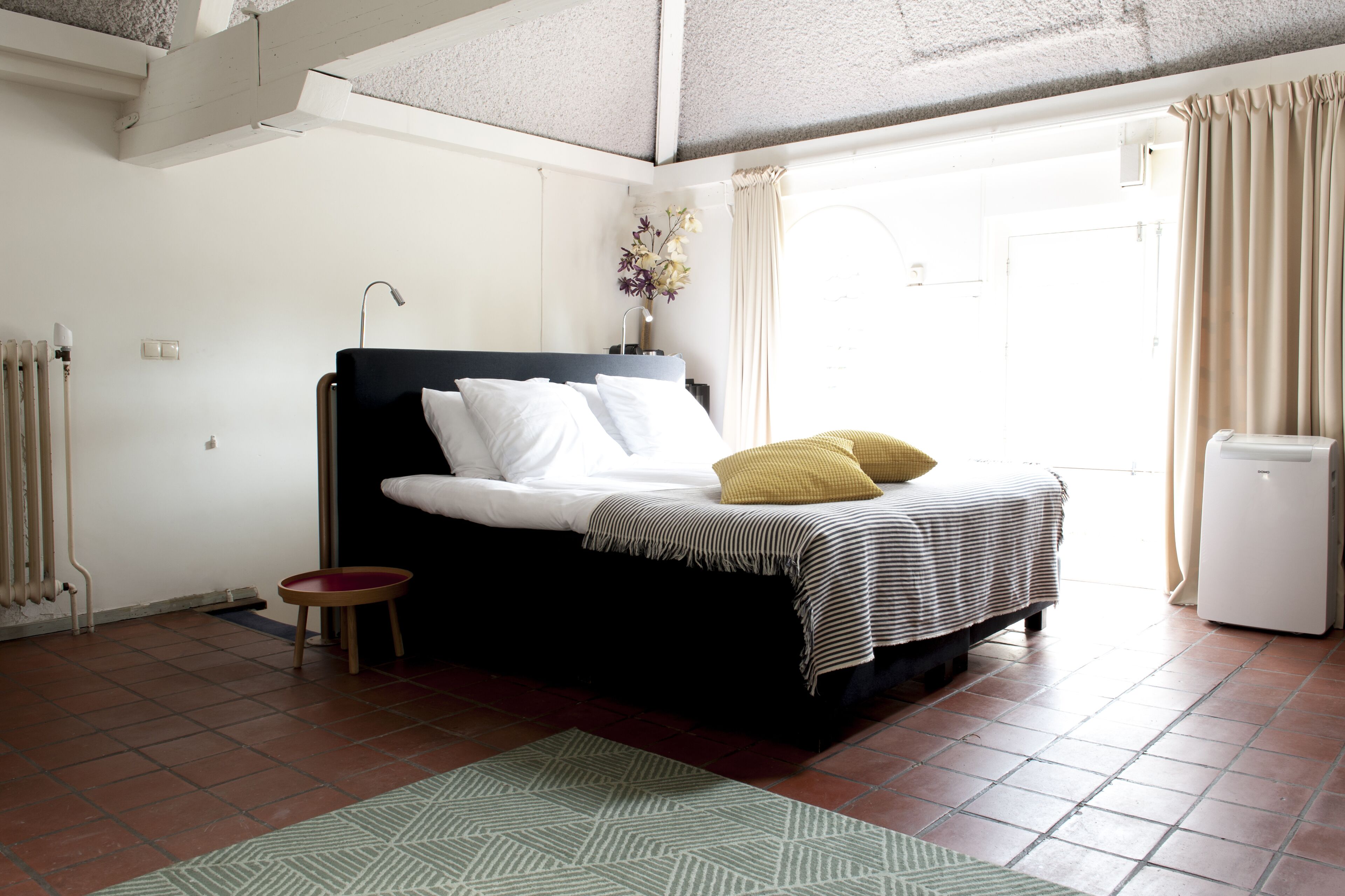 Quarto com cama, mesa de cabeceira pequena, radiador e janela com cortinas.