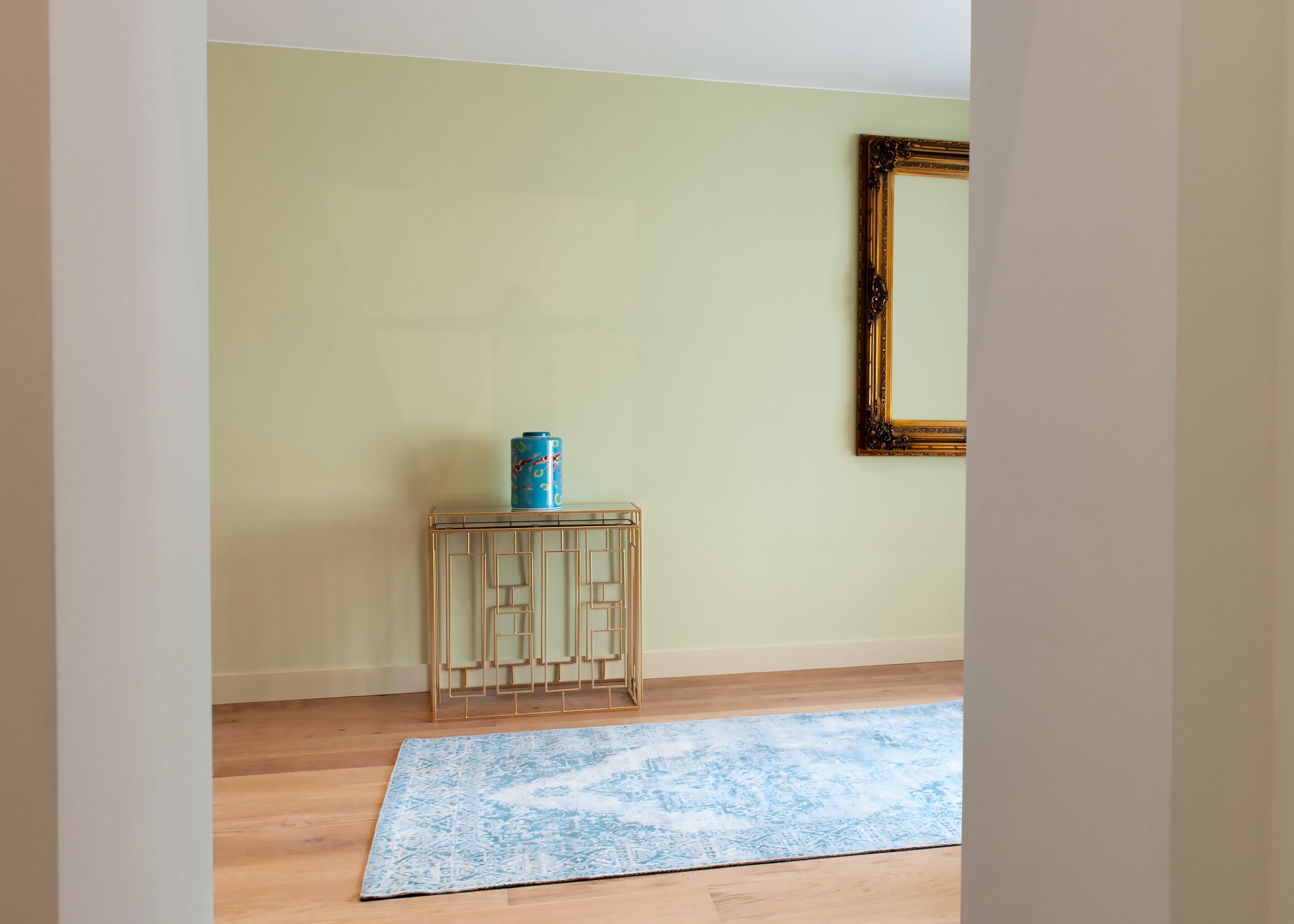 Una habitación con pared verde claro, suelo de madera, alfombra azul estampada, una pequeña mesita de madera con una lata azul encima y un espejo con marco dorado en la pared.