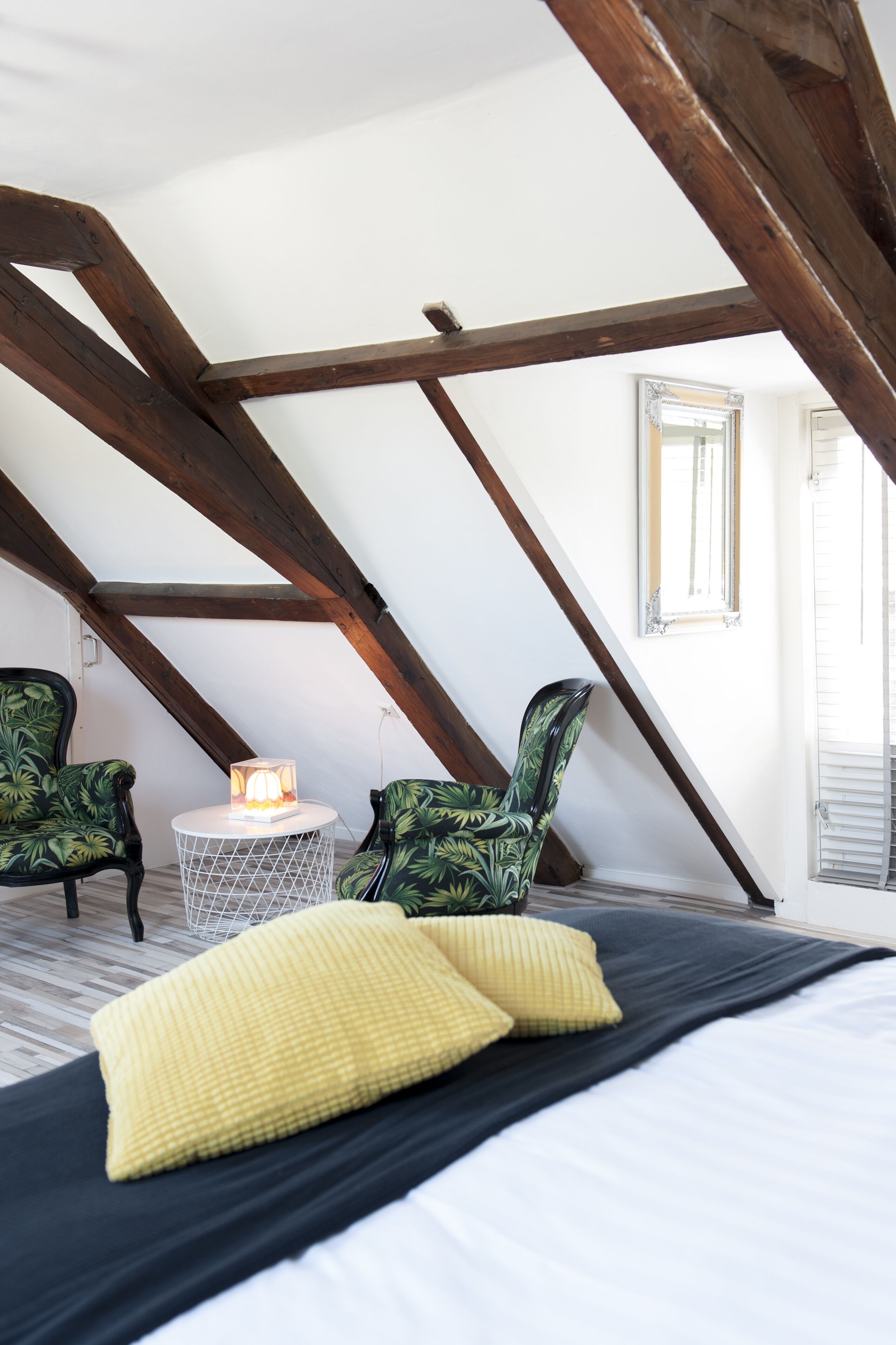 Ein Schlafzimmer mit schrägverdeckter Decke, Holzbalken, einem Bett mit gelben Kissen, zwei grün gemusterten Stühlen und einem kleinen runden Tisch mit einer Lampe.